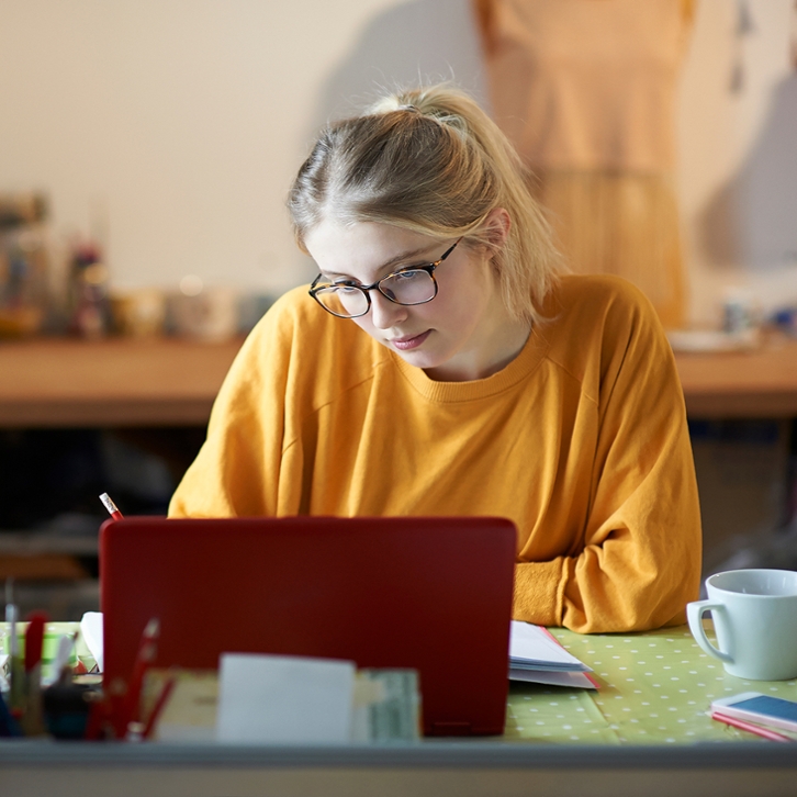 Kobieta w pomrańczowej bluzie pracuje przy biurku na laptopie i robi notatki przygotowując zgłoszenie reklamacyjne.