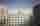 Dziedziniec nowoczesnego kompleksu budynków o jasnych elewacjach i licznych oknach. Pośrodku widoczna jest flaga Unii Europejskiej na maszcie, a nad budynkami rozciąga się pochmurne niebo.